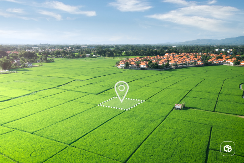 Vista aérea de uma extensa área agrícola com plantações verdes organizadas em grandes lotes geométricos. No centro da imagem, há uma marcação gráfica destacando um terreno específico, representado por um contorno pontilhado e um ícone de localização. Ao fundo, é possível ver um conjunto residencial, árvores, montanhas distantes e céu parcialmente nublado. A imagem remete a propriedade rural, investimento em terras, agronegócio, georreferenciamento e valorização imobiliária.
