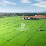 Vista aérea de uma extensa área agrícola com plantações verdes organizadas em grandes lotes geométricos. No centro da imagem, há uma marcação gráfica destacando um terreno específico, representado por um contorno pontilhado e um ícone de localização. Ao fundo, é possível ver um conjunto residencial, árvores, montanhas distantes e céu parcialmente nublado. A imagem remete a propriedade rural, investimento em terras, agronegócio, georreferenciamento e valorização imobiliária.